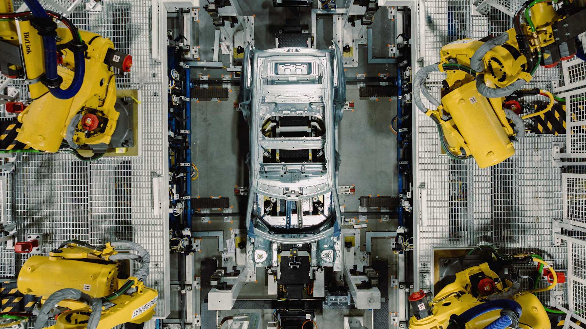 Overhead shot shows a car chassis being assembled by four yellow robotic arms in a factory. The chassis is silver and surrounded by a metal framework.