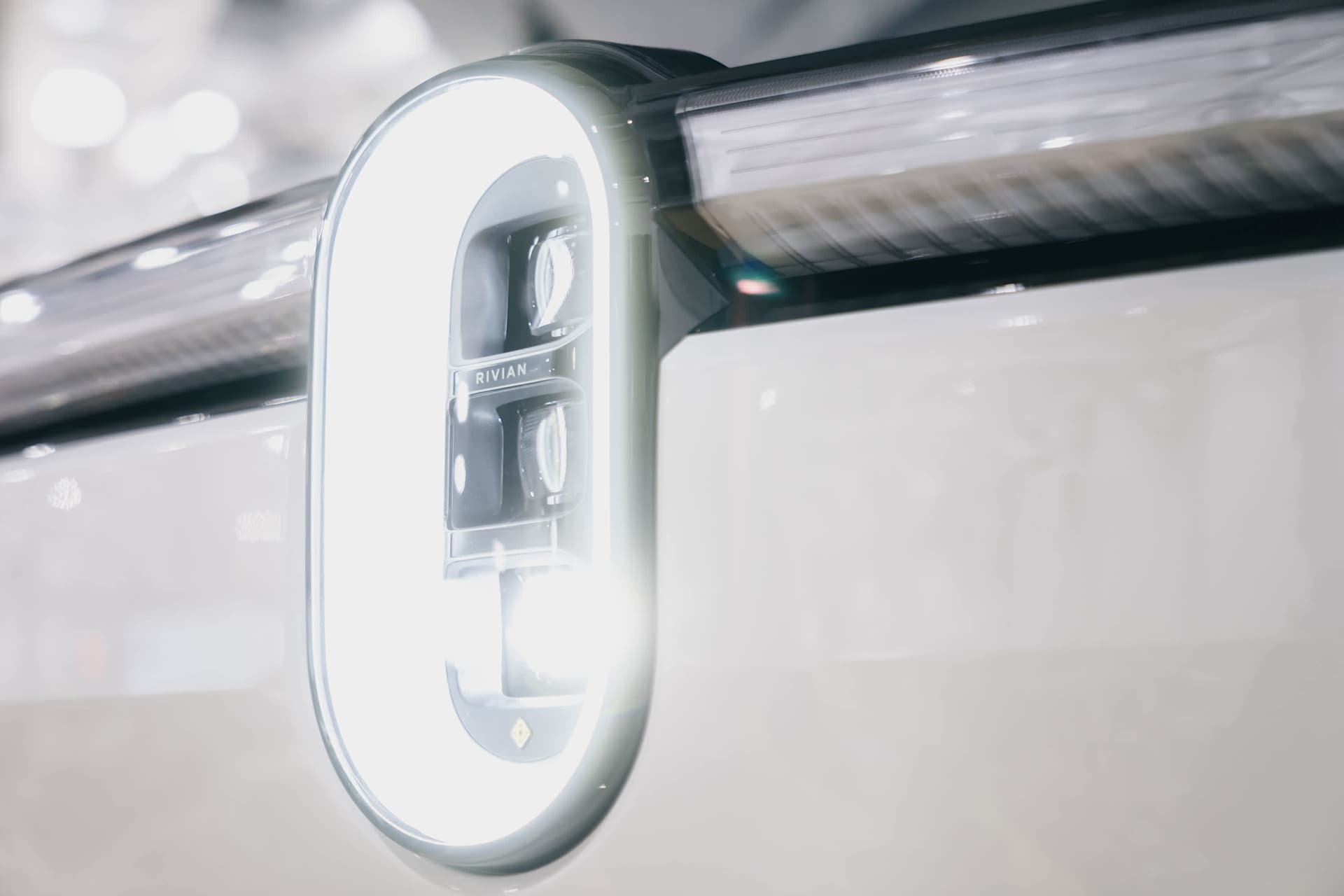 Close-up of a Rivian vehicle's vertical headlight. The light is encased in an oval-shaped frame, emitting a bright glow. "Rivian" is visible on the li