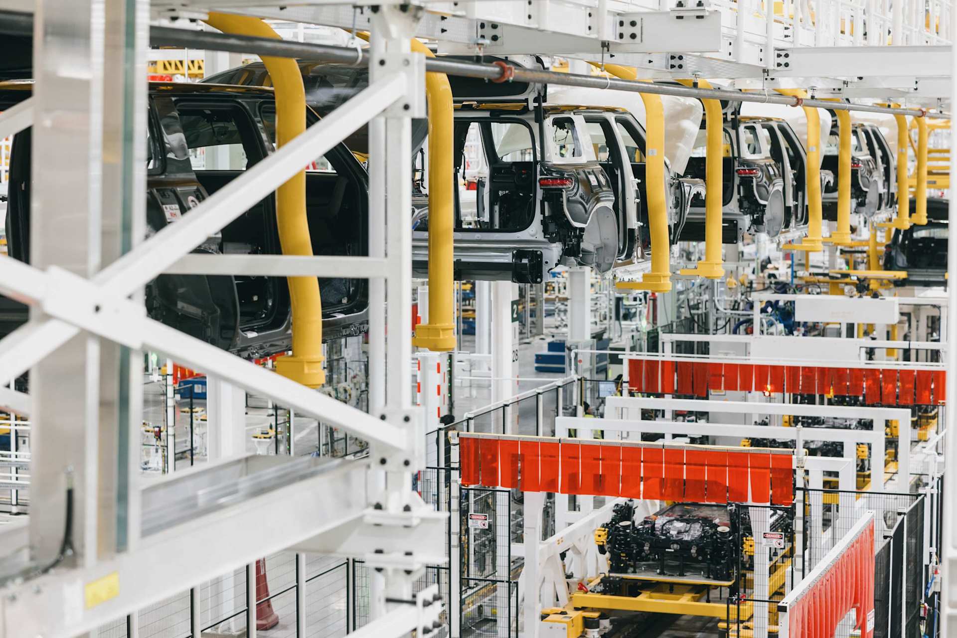 Car bodies hang from yellow supports along a white factory assembly line. Below, auto parts move on a parallel track, ready for installation.