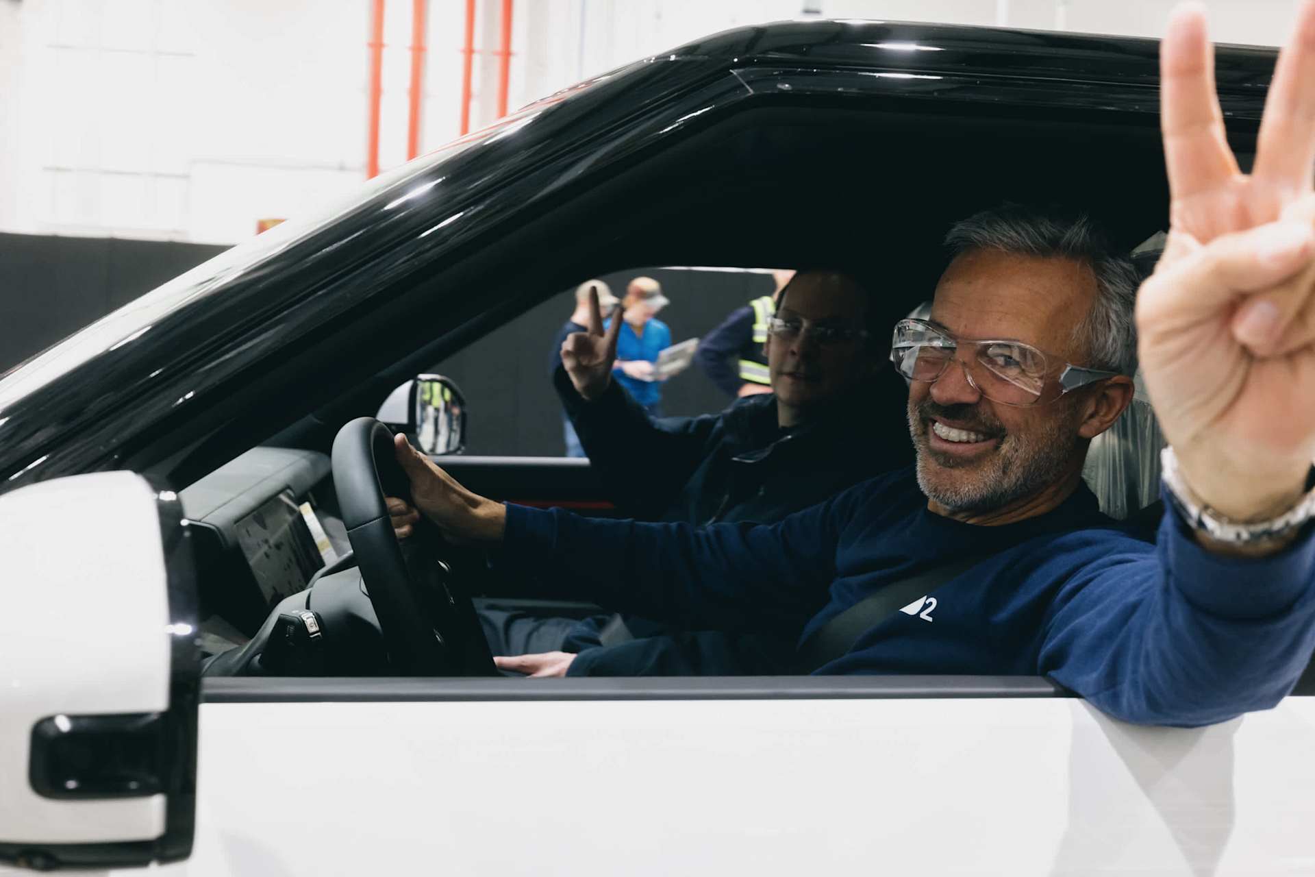 Man in driver's seat flashes peace sign, smiling. He wears safety glasses and a blue shirt with a logo. Passenger also flashes peace sign.