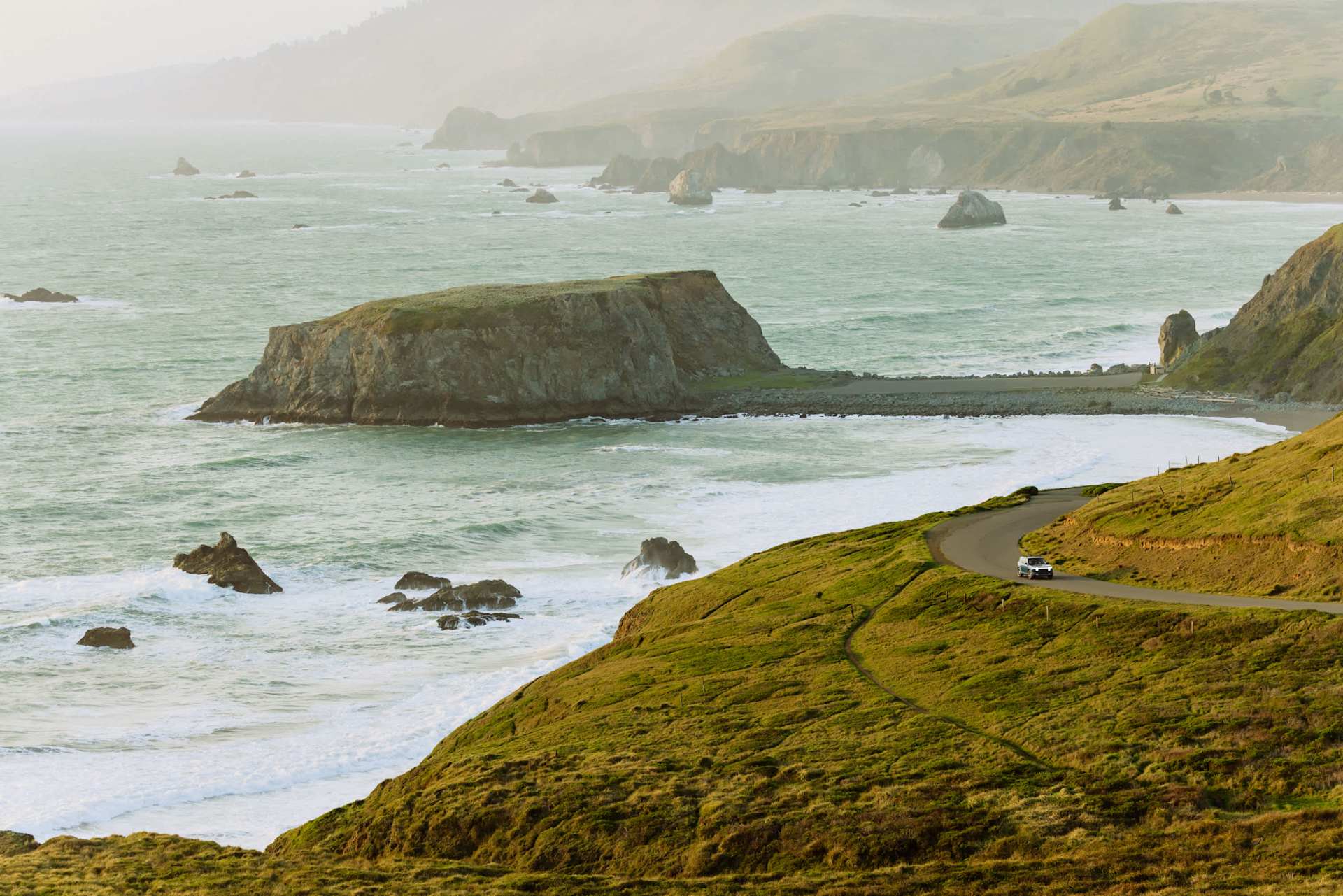 Coastal view of a car driving on a winding road along a grassy hillside. Ocean waves crash against rock formations in the water. Hazy sunlight.