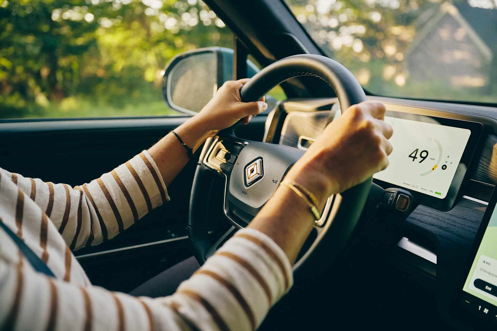 A person drives a Rivian; the dashboard screen reads 49 mph. The driver wears a striped shirt and bracelets. Green trees are visible through the winds