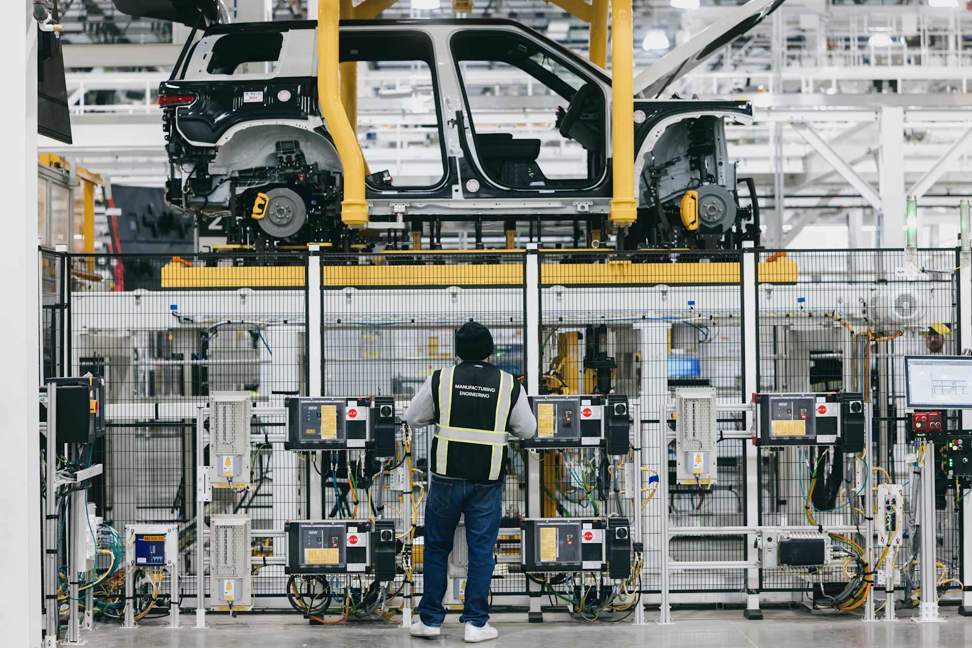 Rivian engineer in safety vest stands in factory, facing a partially assembled SUV body on a yellow platform. The vehicle frame is white and black, wi