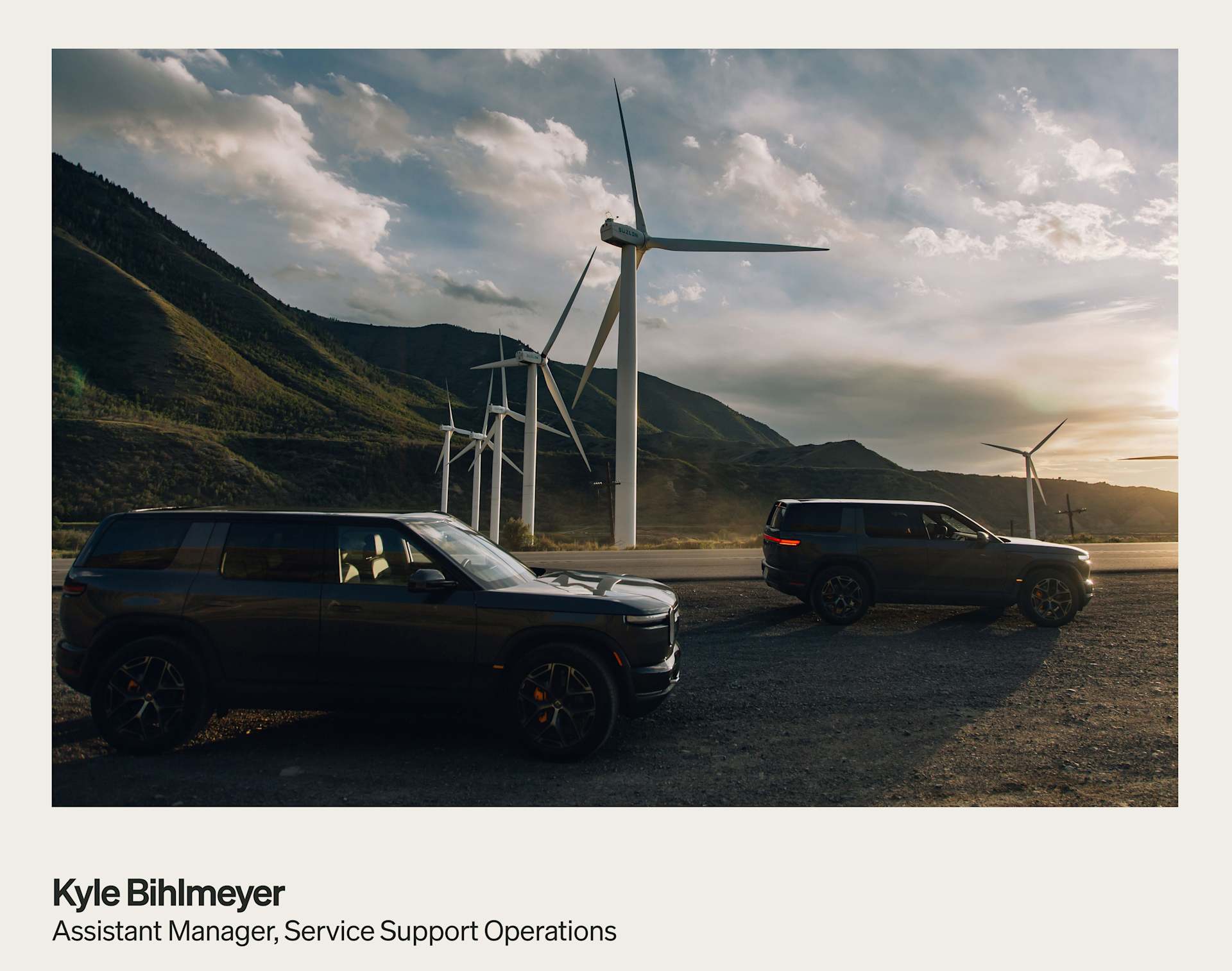 Two Rivian SUVs park on a gravel shoulder with wind turbines and mountains in the background. The sun sets behind the turbines, casting a warm glow.