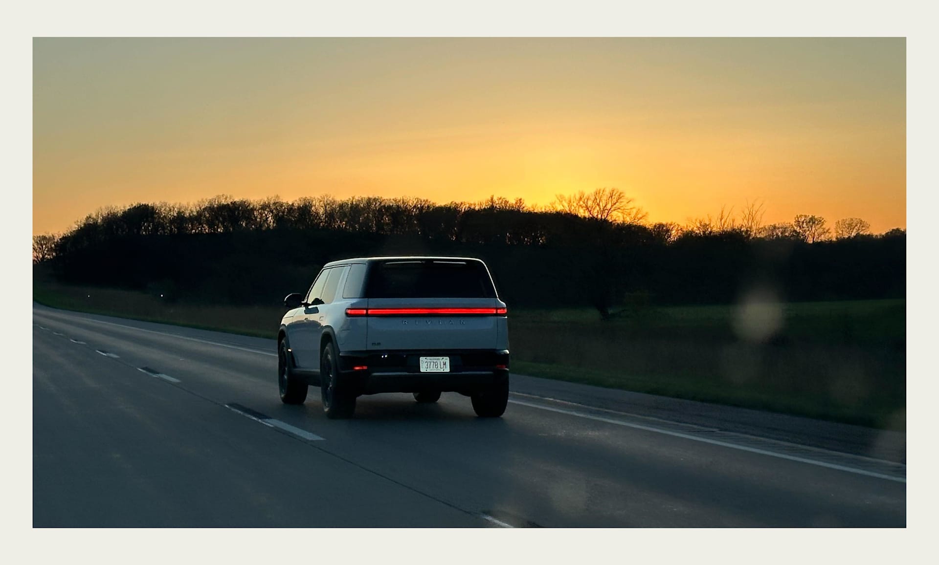 Rivian SUV drives on a highway at sunset. Golden sky behind a dark treeline. Taillights glow on the light gray vehicle.