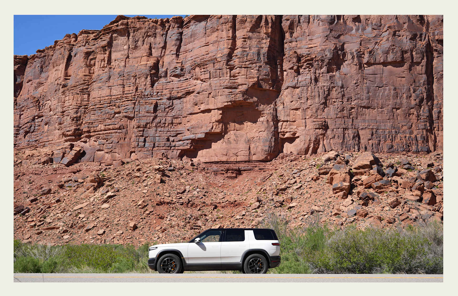 White SUV drives along a road with a large red rock formation in the background. Green bushes grow along the roadside under a clear blue sky.