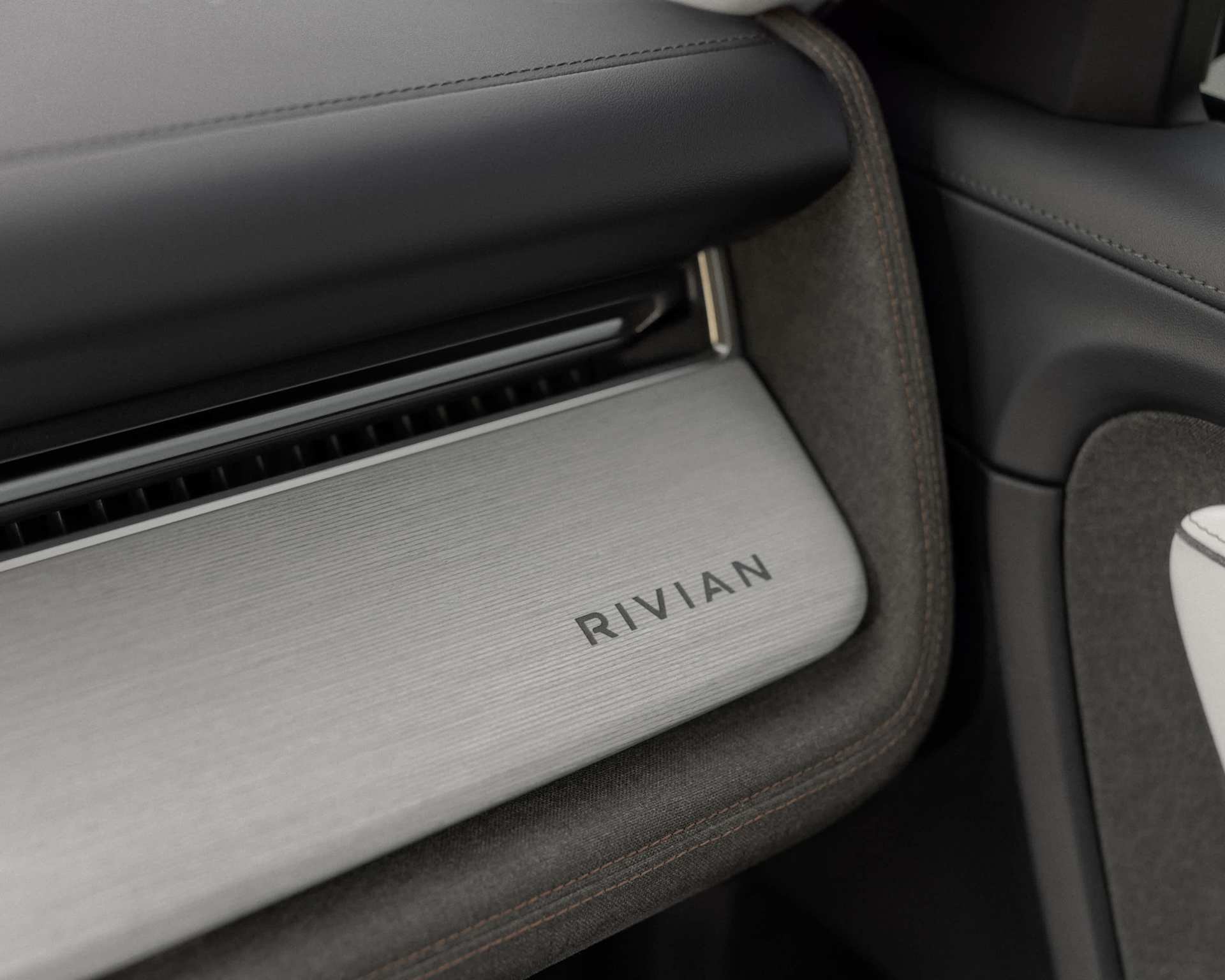 Close-up of the Rivian dashboard featuring a silver panel with the company name etched on it. The interior is lined with dark gray fabric and black le