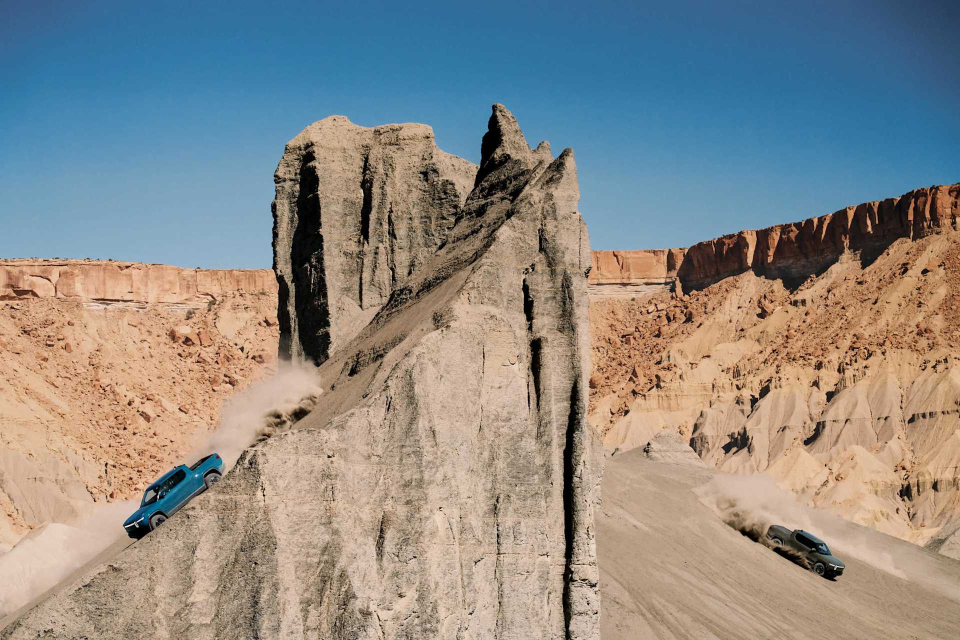 Two Rivian trucks drive down a steep, sandy hill, kicking up dust. A tall, gray rock formation rises in the center against a clear blue sky. Orange cl