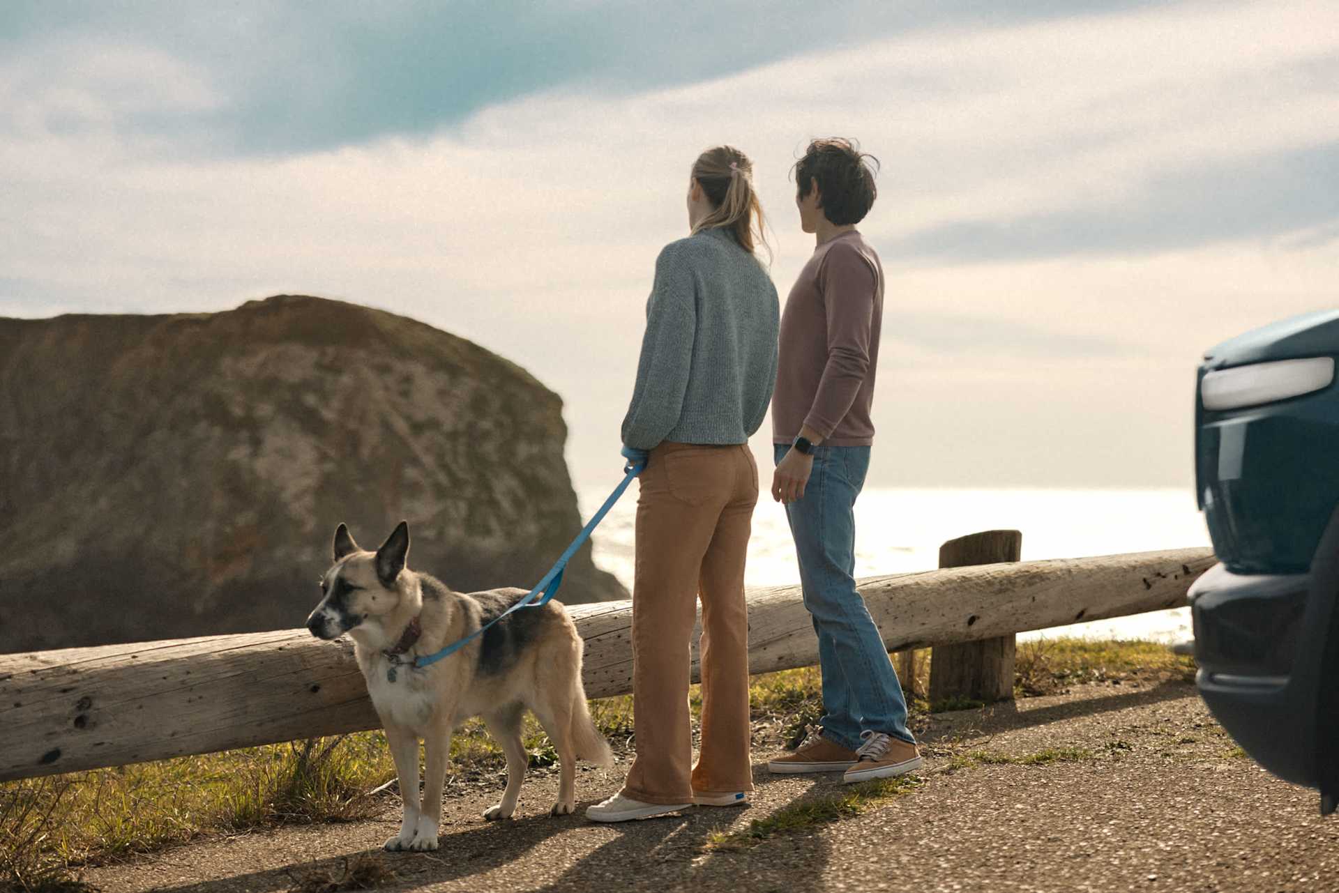 Couple and dog stand on a coastal path, looking out at the ocean. A blue truck is parked nearby. The scene evokes a sense of peaceful exploration.