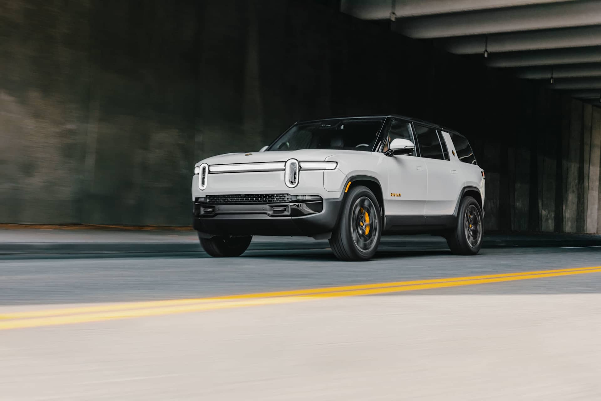 Rivian SUV drives on a road through a concrete tunnel. The vehicle is light gray with black trim and yellow brake calipers. Motion blur indicates move