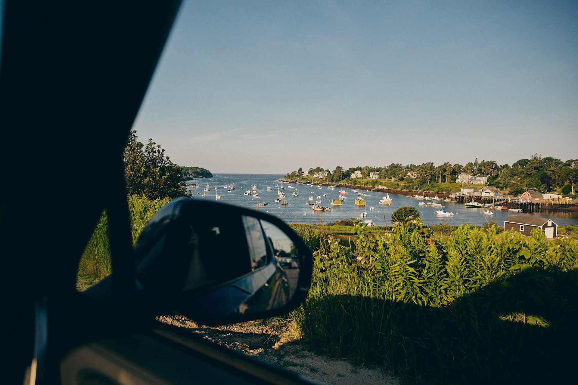 Car side mirror reflects a harbor dotted with sailboats. Lush green foliage lines the shore under a clear blue sky, seen from the car window.