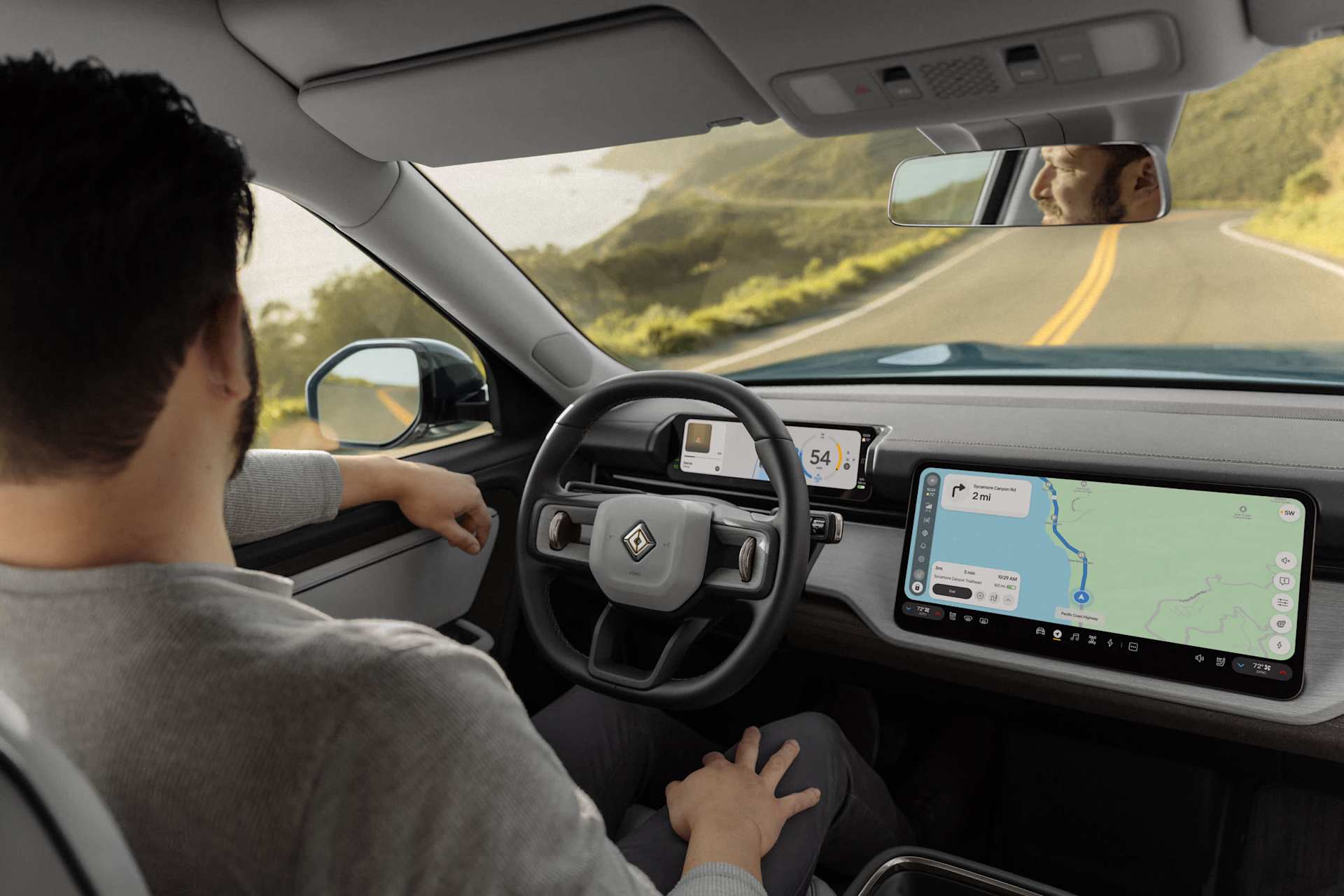 A man rides in a self-driving car, dashboard displays navigation. The car travels on a coastal road, seen through the windshield and rearview mirror.