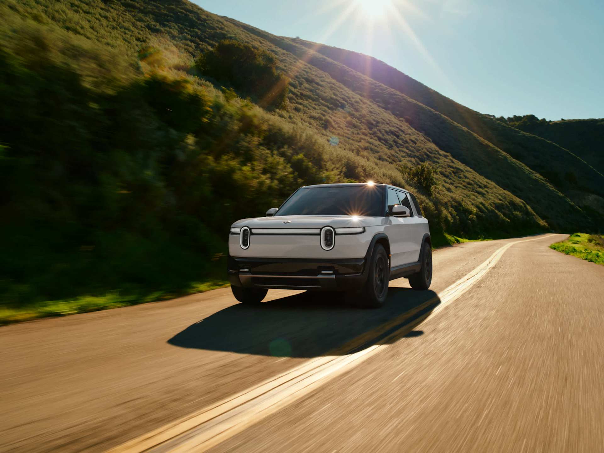 Rivian EV truck drives on a winding road through green hills, sun shining overhead. The truck is light gray with black trim.