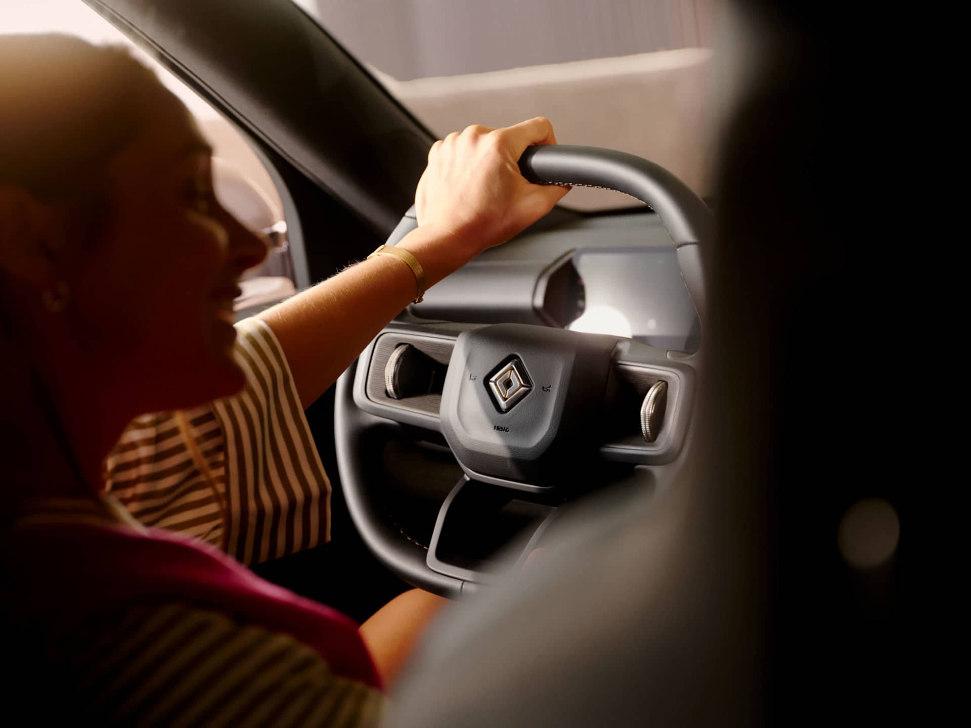 A woman drives a Rivian EV, hand on the steering wheel, Rivian logo visible. The interior is dark, with sunlight glinting off the dashboard.