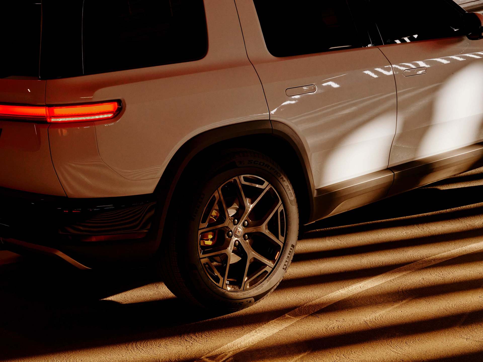 Rear quarter view of a white Rivian SUV parked on a textured surface, casting strong linear shadows. Taillight glows red, highlighting the vehicle's m