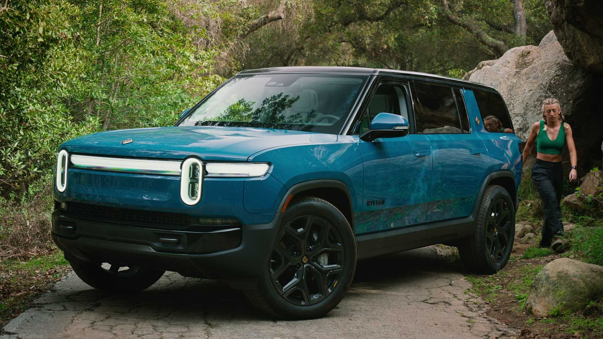 Rivian electric SUV parked on a dirt road in a wooded area. A woman in athletic wear walks beside the vehicle.