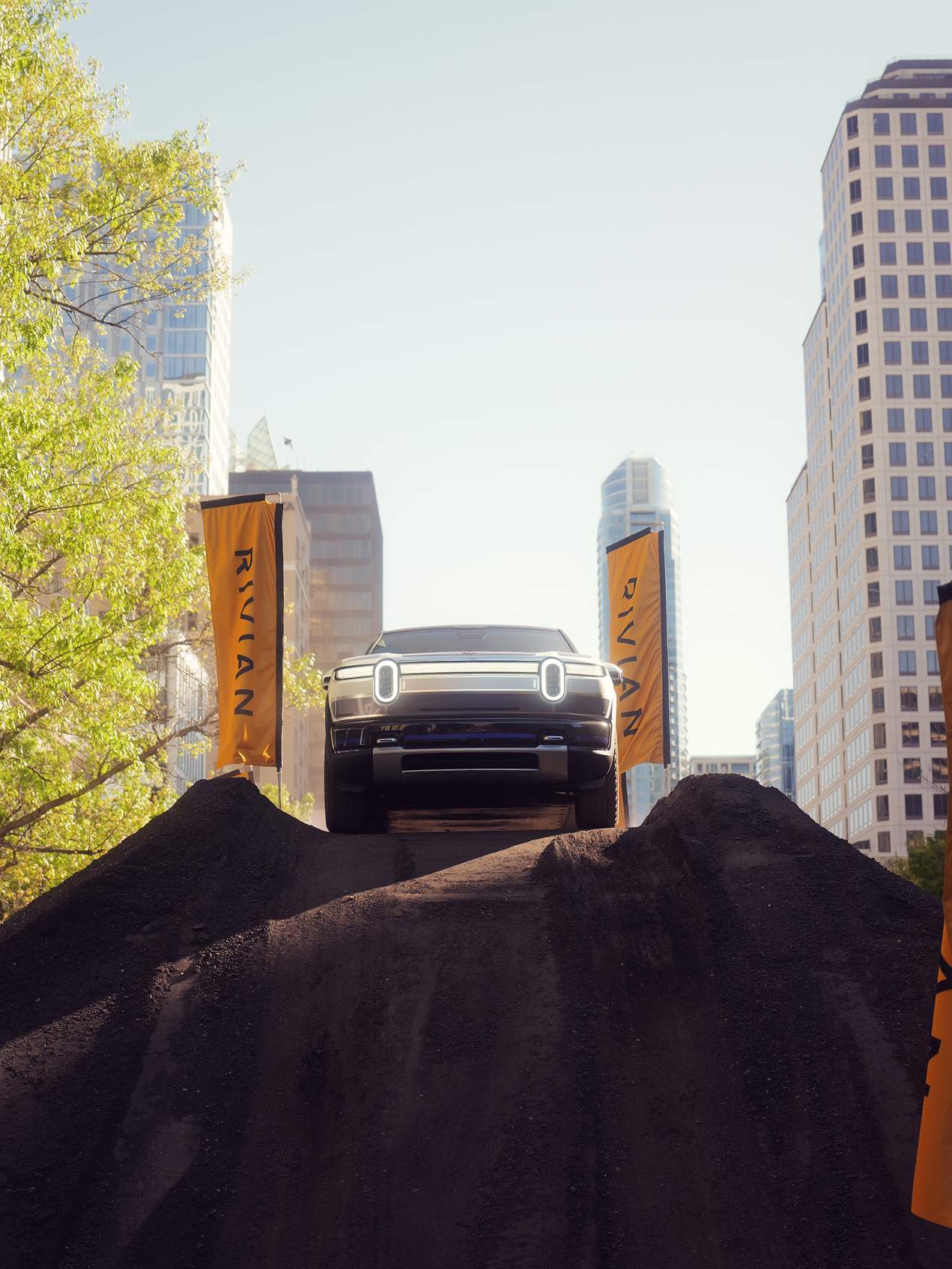 Rivian electric truck ascends a dirt hill, front view, in an urban setting. "Rivian" banners flank the vehicle against a backdrop of tall buildings, d