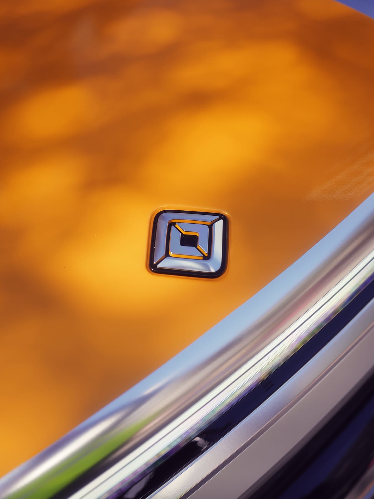 Close-up of a Rivian logo on the hood of an orange vehicle, with a chrome accent.