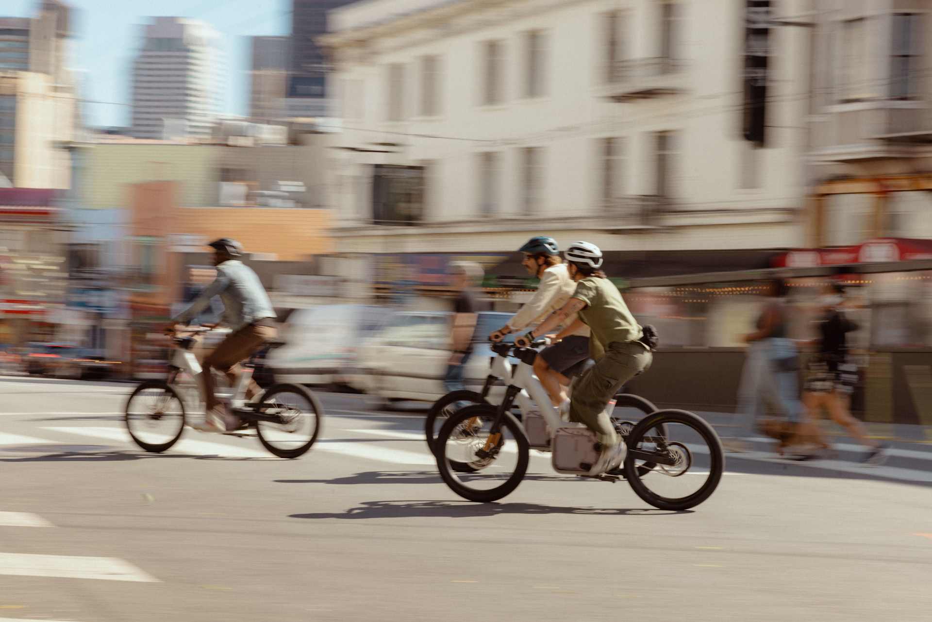 Three people ride Rivian e-bikes across a city street. The cyclists wear helmets. Buildings and blurred traffic line the background. The image uses mo