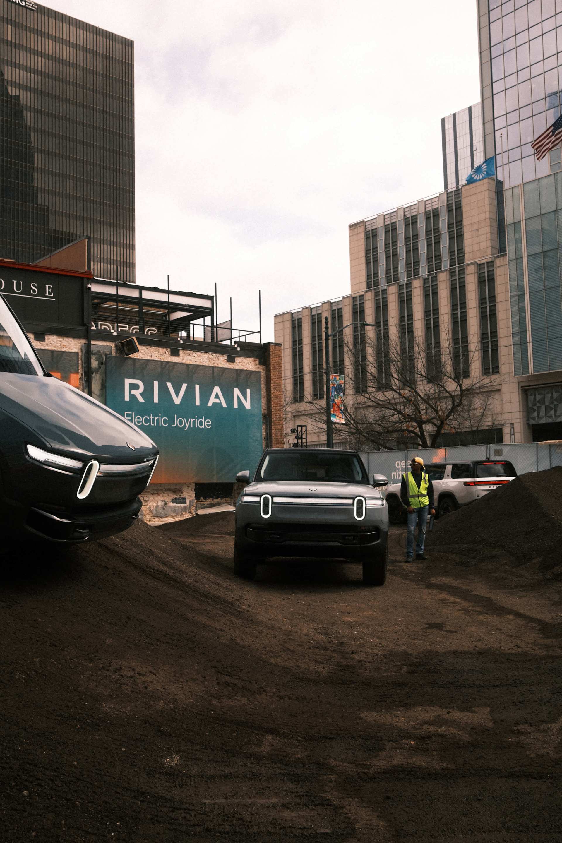 Two Rivian electric vehicles sit on a dirt track in an urban setting. A banner reads "RIVIAN Electric Joyride." A worker in a safety vest stands nearb