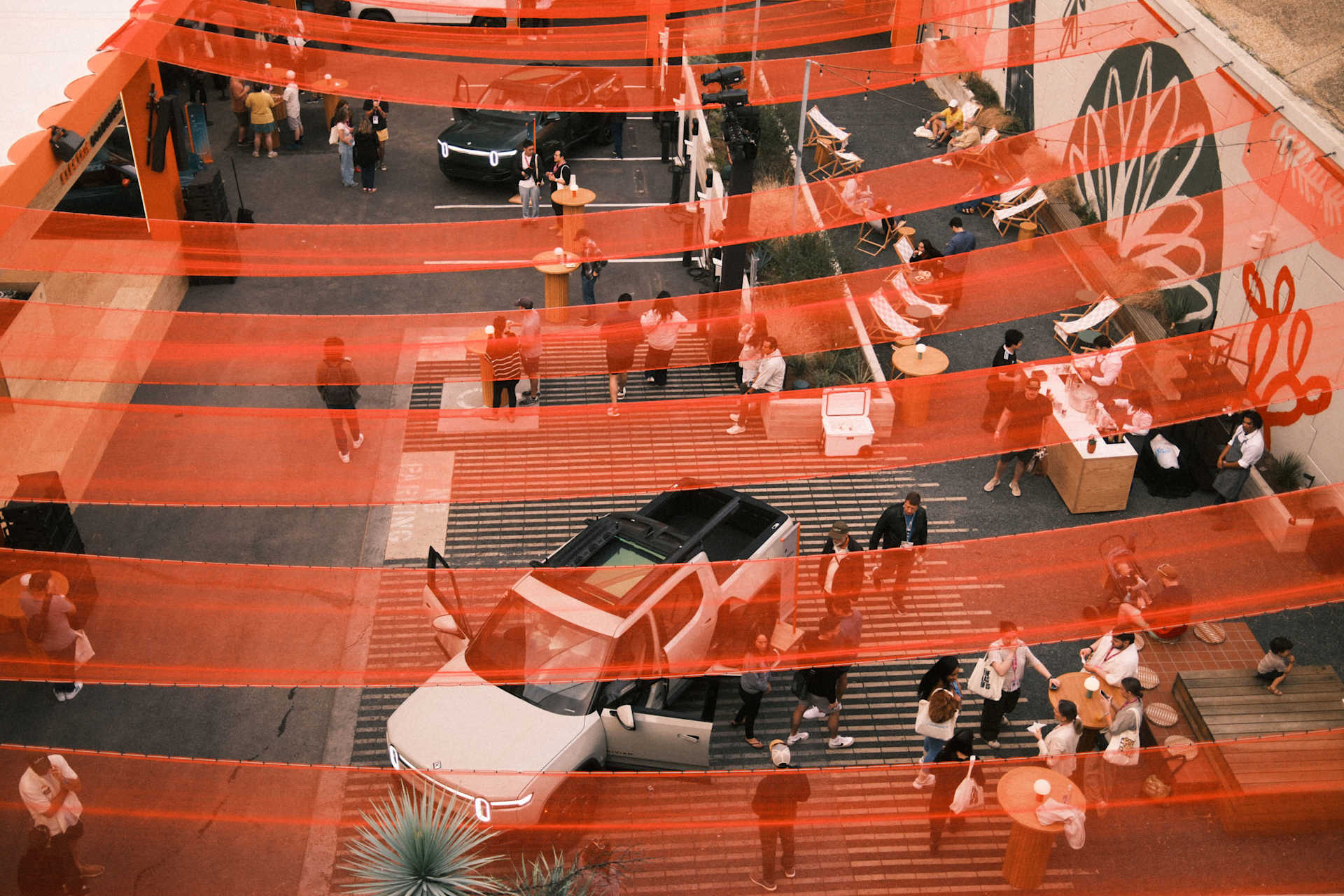 Overhead view of Rivian vehicles on display at SXSW, surrounded by people. Orange mesh netting hangs overhead. Some people are seated in lounge chairs
