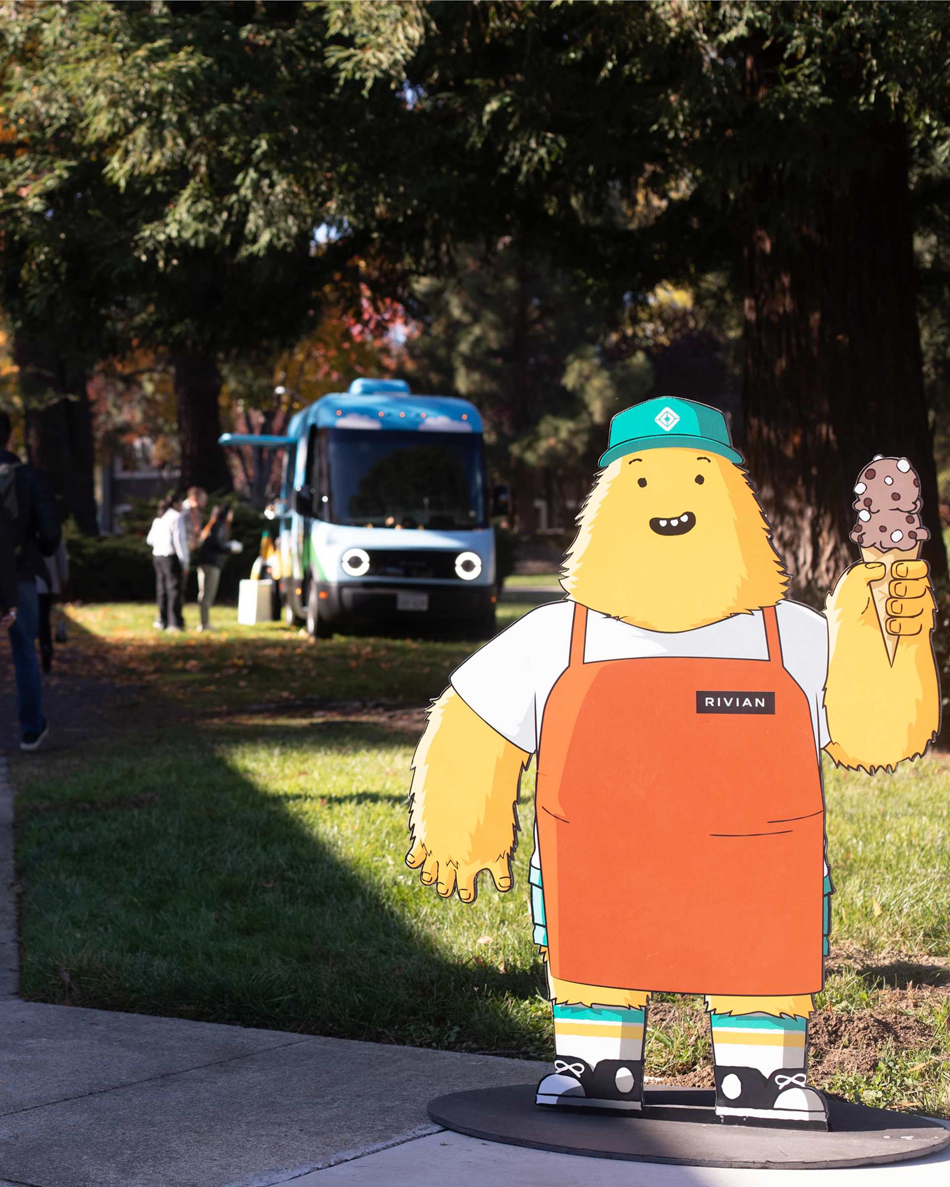 Cartoon cutout of a fuzzy, yellow Rivian mascot holds an ice cream cone. Mascot wears a green hat, orange apron, and striped socks. A Rivian truck is 