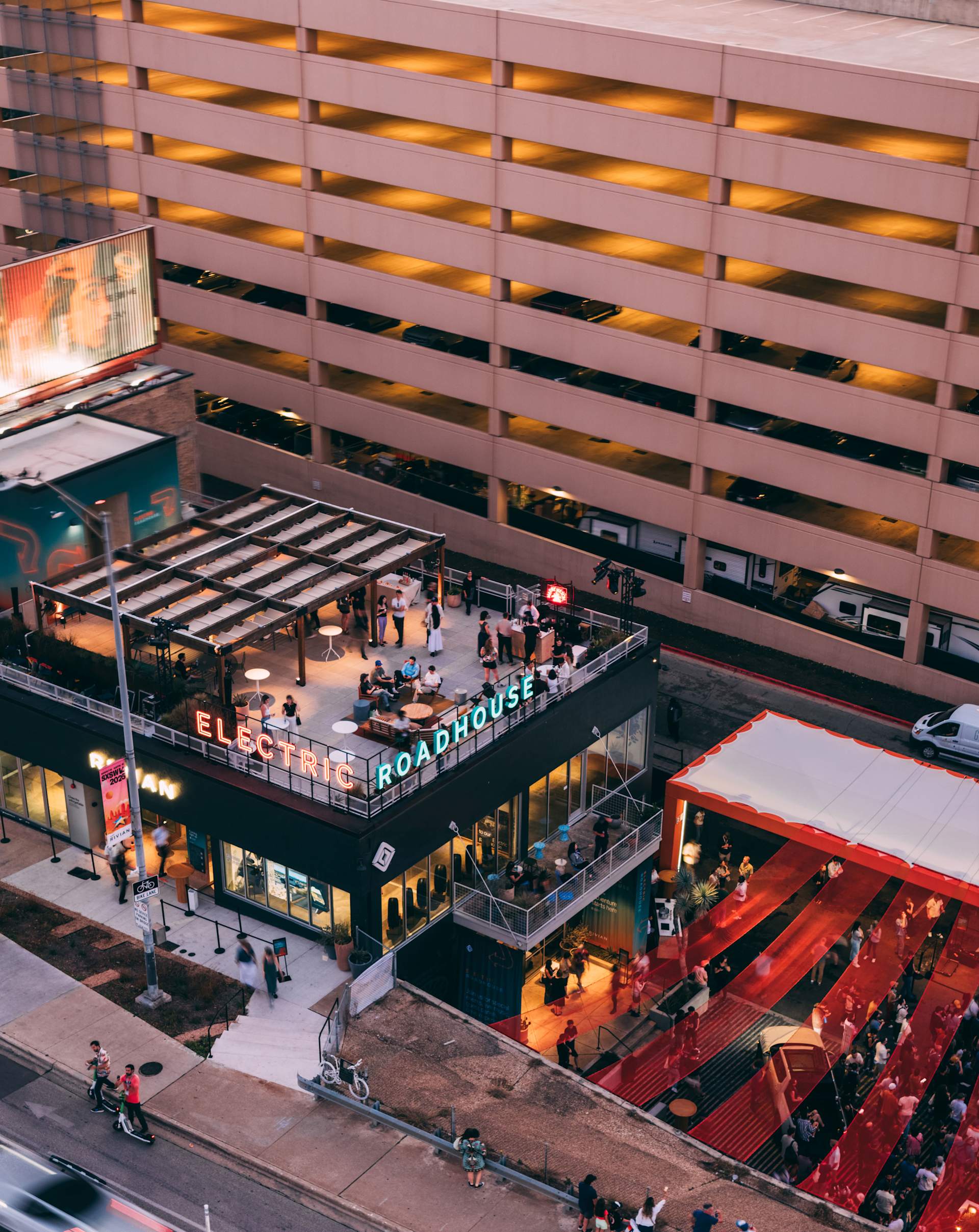 Aerial view of Rivian's "Electric Roadhouse" at SXSW. People gather on the rooftop under a pergola. Neon sign reads "Electric Roadhouse". A red-carpet
