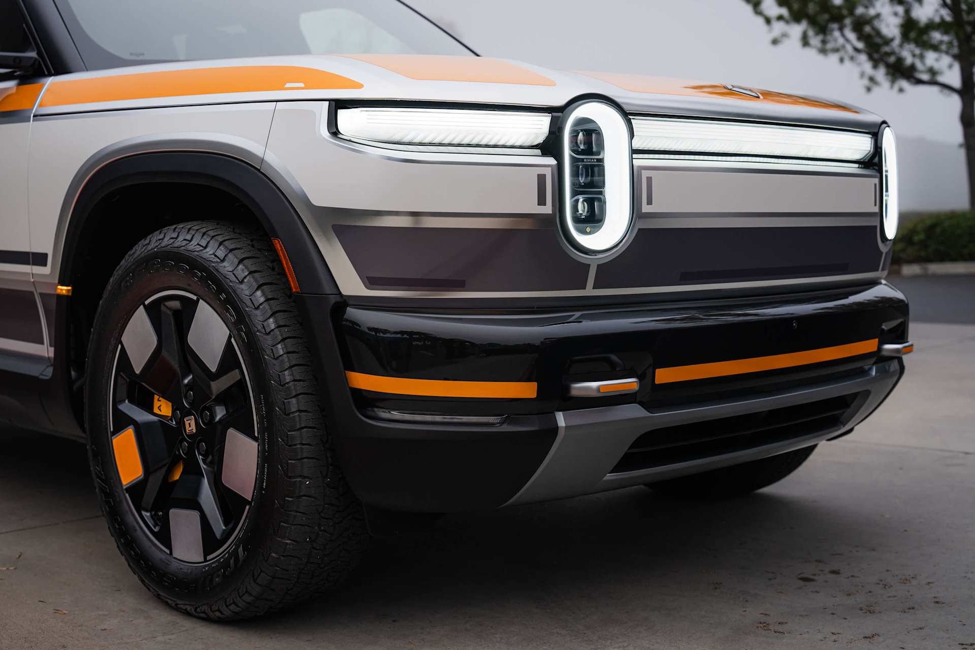 Rivian truck front view shows custom paint job with orange stripes and accents. Headlights are illuminated, creating a distinctive oval shape.