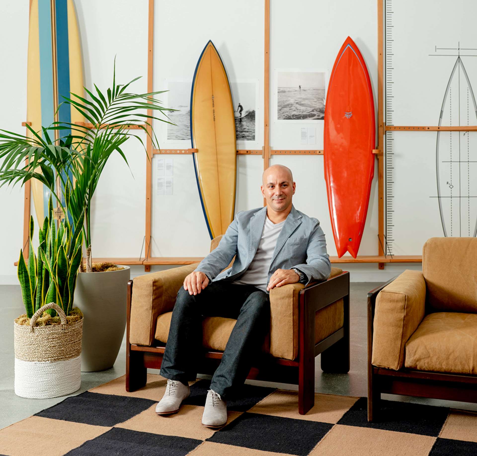 A man sits in a chair in front of surfboards. The bald man wears a gray jacket and dark jeans. Behind him, three surfboards hang on a white wall, alon
