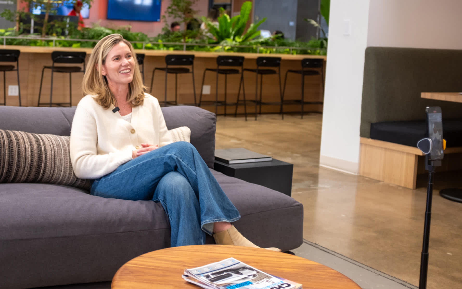Woman sits on a gray couch, smiling, in a modern office space. A phone on a tripod stands in front of her. A magazine rests on a round wooden table.