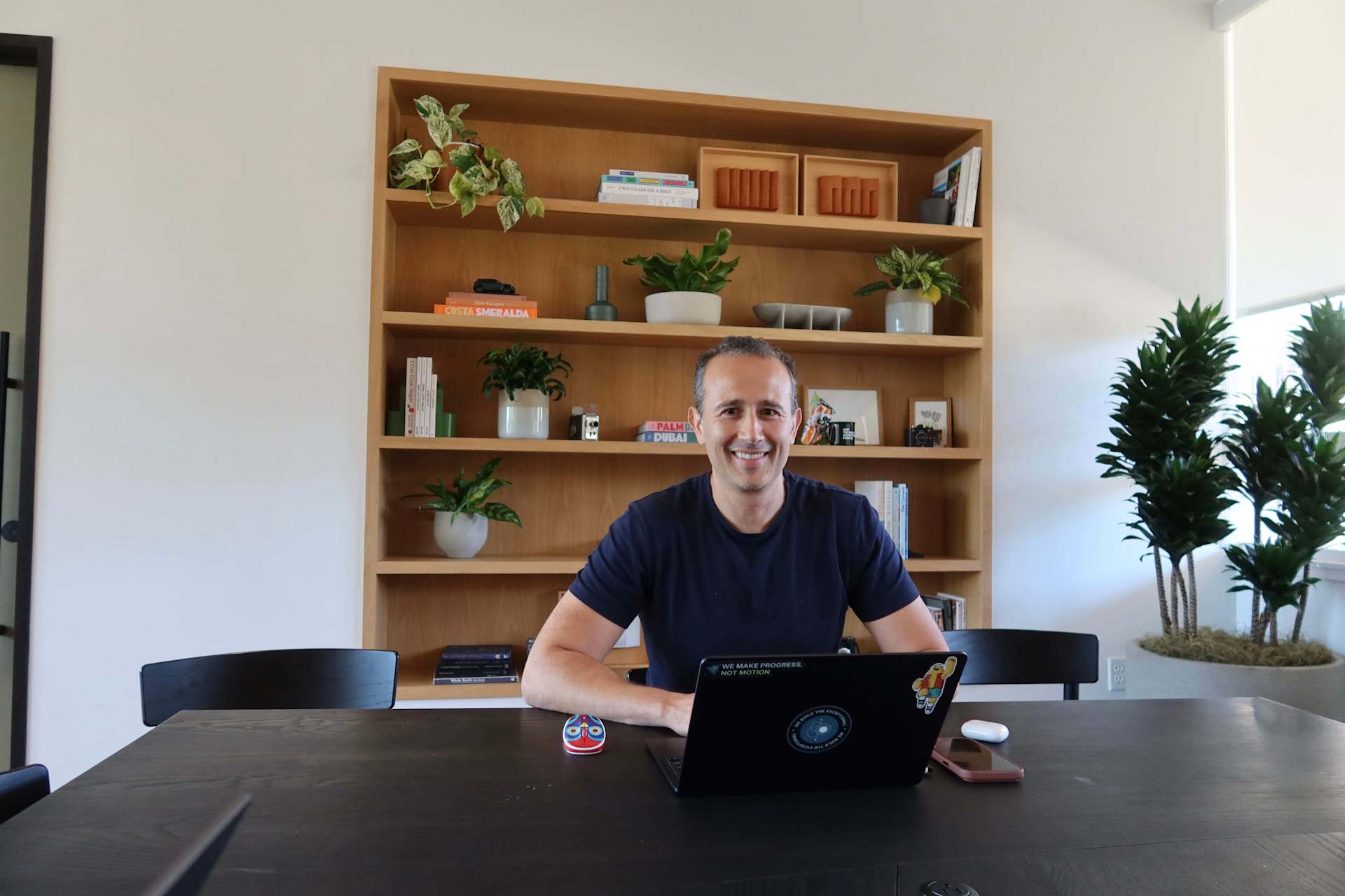Wassym Bensaid sits at a desk, smiling. Behind him is a bookshelf filled with plants and books. His laptop reads, "We make progress, not motion."