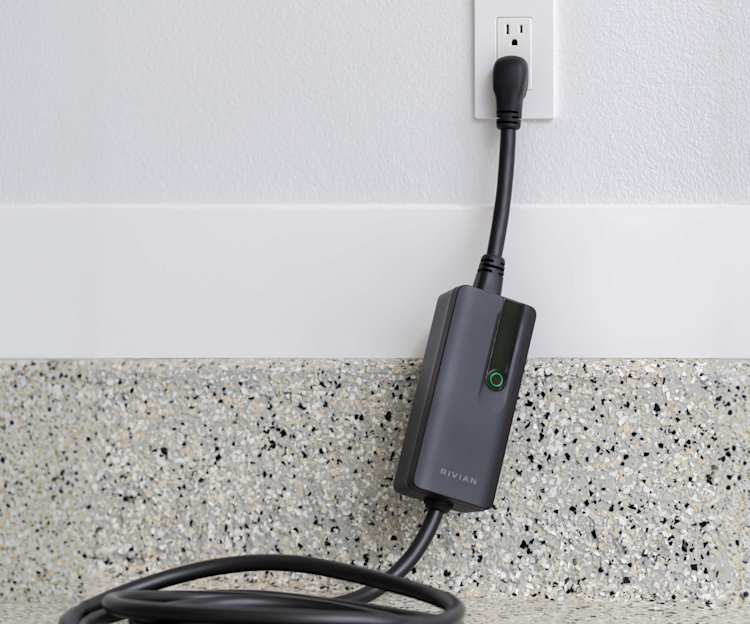 Rivian EV charger plugs into a wall outlet above a speckled countertop. The black cable is coiled on the counter below the gray charging unit.
