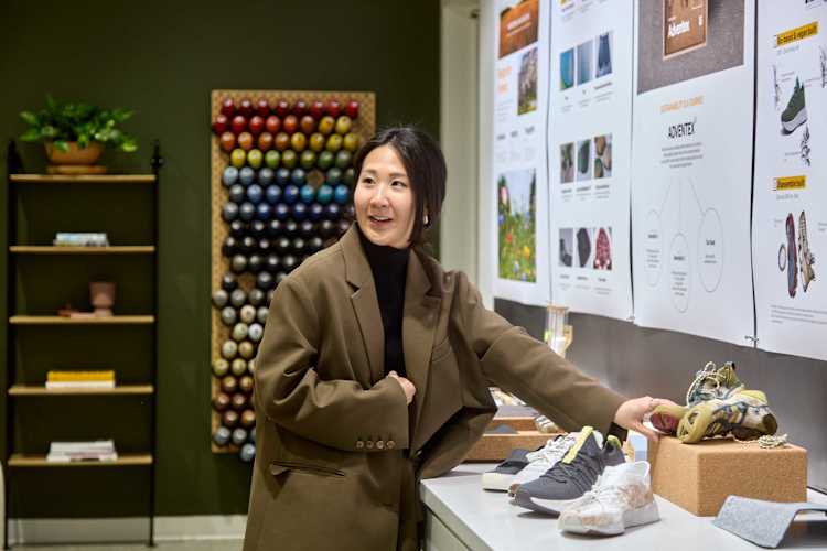 A woman in a brown blazer smiles, displaying sustainable sneakers. Behind her are posters about "Adventex" and a wall of colorful yarn spools.