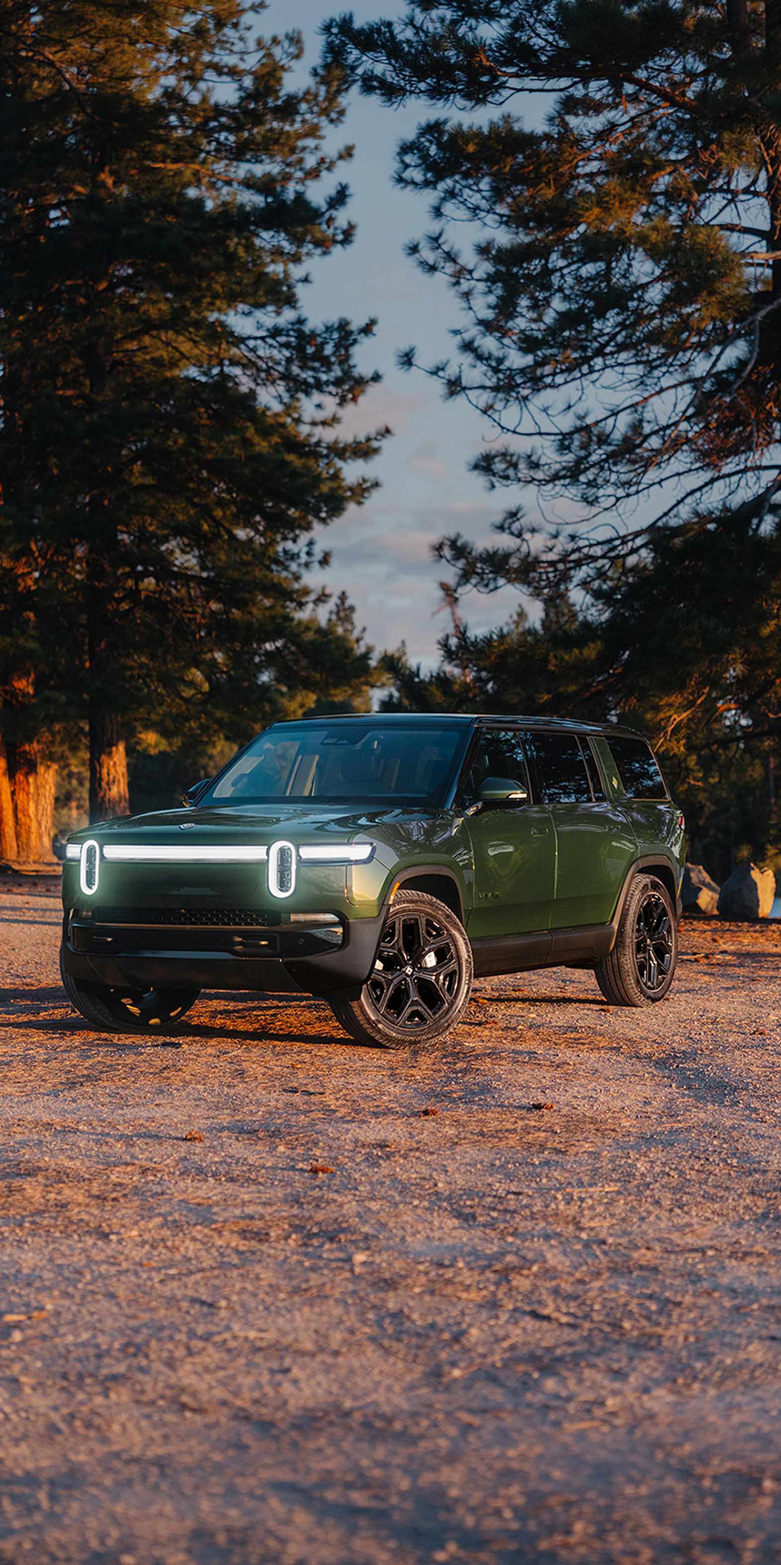 orest Green Rivian R1S electric SUV parked on a dirt path near a lake surrounded by pine trees.